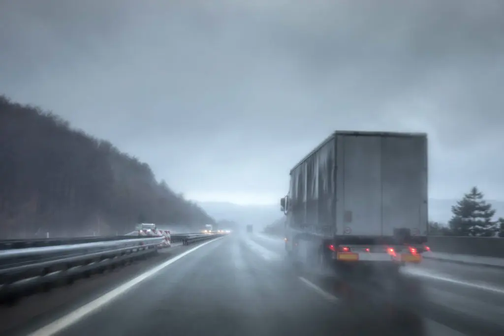 Semi-truck driving in bad weather conditions on rain-soaked road.